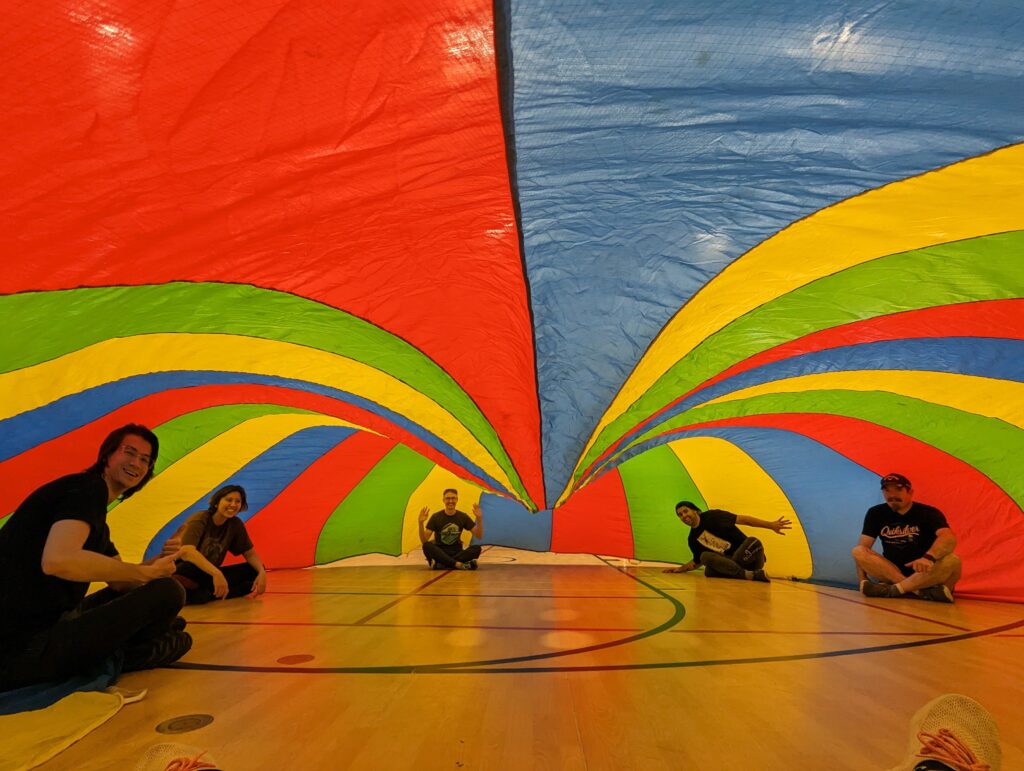 A picture of several folks underneath a brightly coloured parachute inside of a gym. They're all smiling at the camera. 