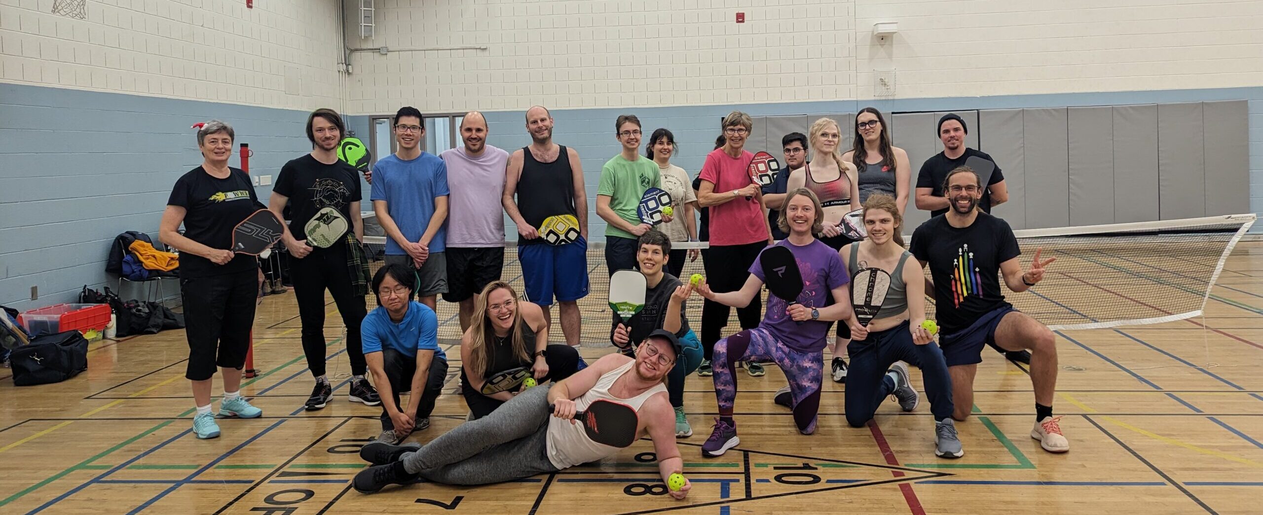 A group of about 20 folks are standing in a gym in athletic wear, all holding pickleball paddles and smiling at the camera. This is an image from our Multi-Sport Program!